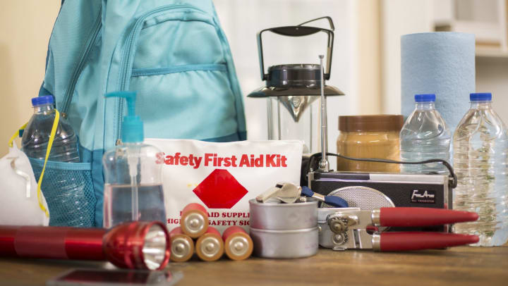 A collection of supplies including flashlight, backpack, batteries, water bottles, first aid kit, lantern, radio, can opener, and mask.