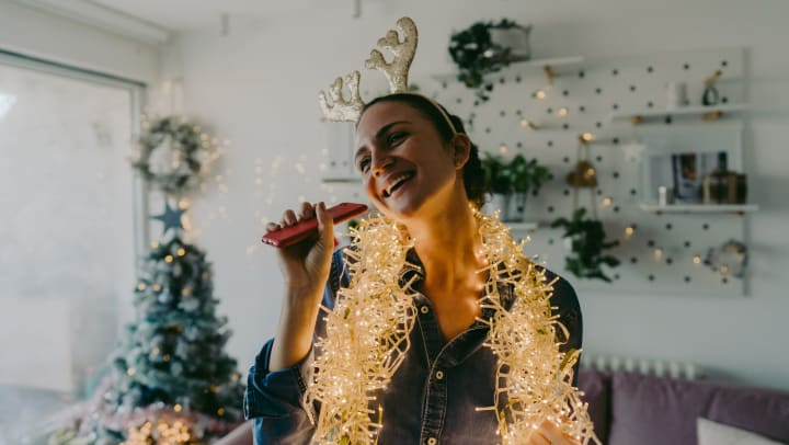 Smiling young woman holding a phone and wearing costume reindeer antlers in a white interior with a tree, wreath, and holiday decorations in the background