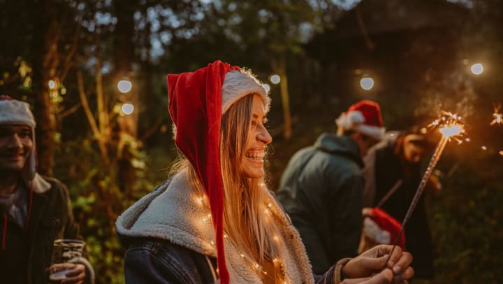 A young, smiling woman wearing a Santa hat outdoors and holding a sparkler, surrounded by smiling people.