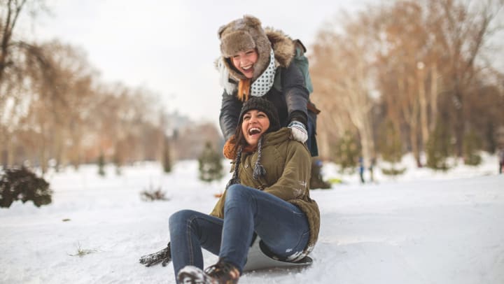 A young woman sitting on a sled and laughing as another woman tries to push her.