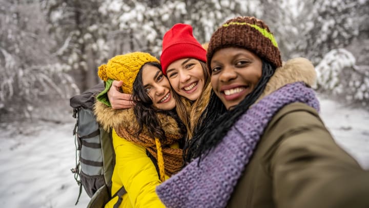 Three smiling young women hug each other as they take a selfie with a snowy nature scene in the background.
