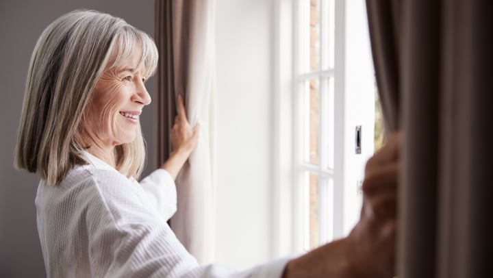 A smiling senior woman opening curtains looks out a bright window.