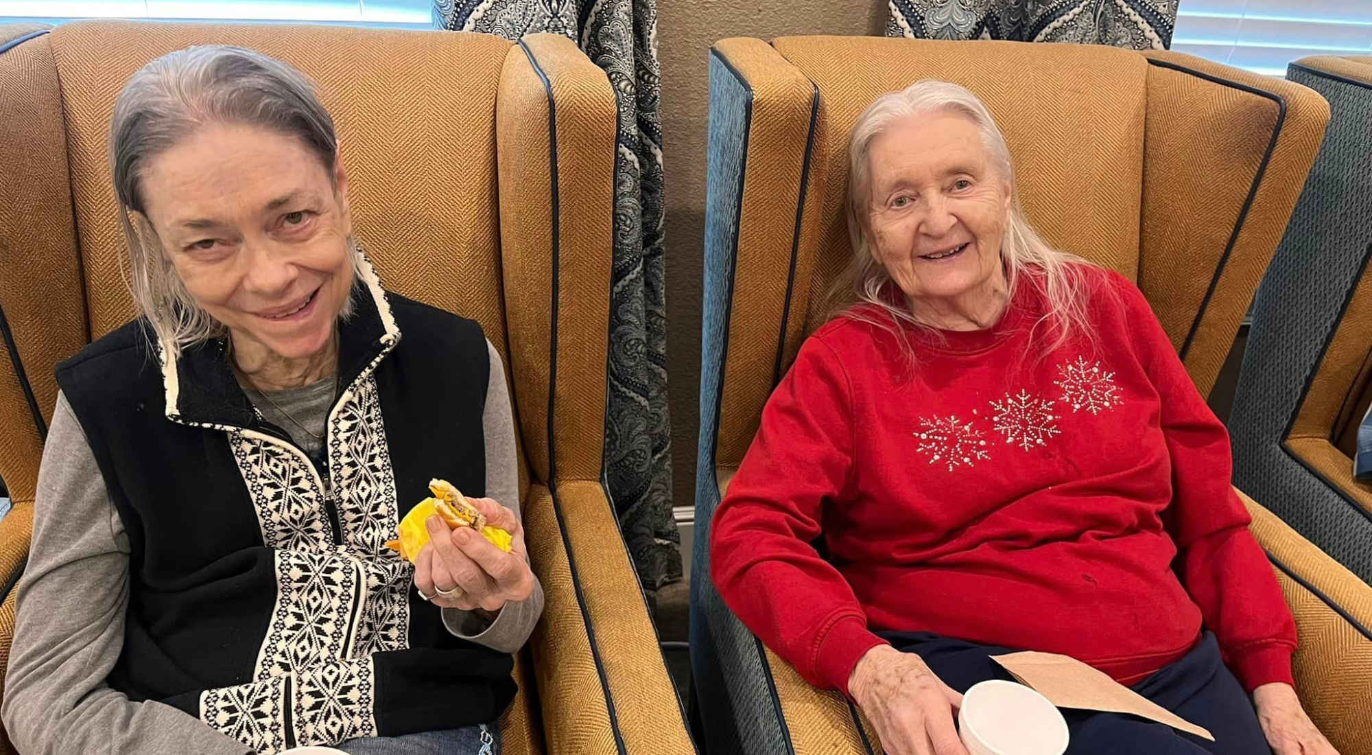 Two senior women relaxing in cozy armchairs with breakfast and coffee.