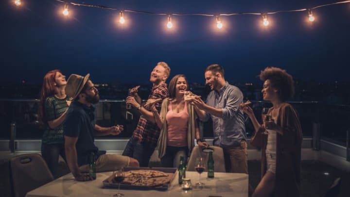 A laughing and smiling group of friends holding beverages and pizza, sit and stand around a table on a balcony, under a string of lights. 