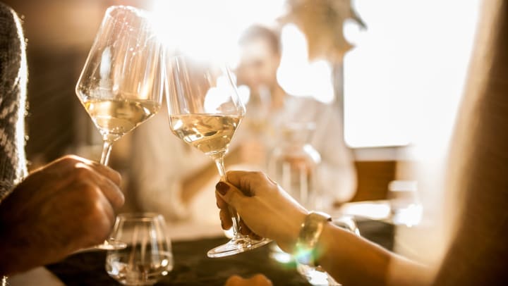Two hands holding two glasses of white wine together in a toast in the foreground with a blurred people in the background. 