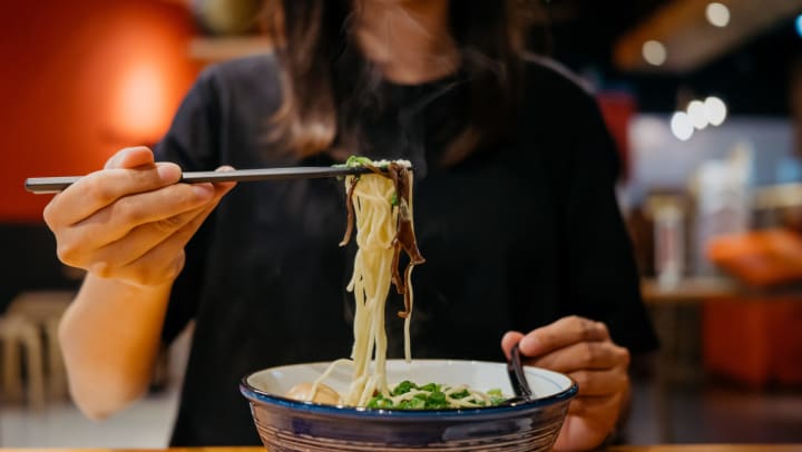 Young woman in a casual restaurant holding chopsticks and eating a hot bowl of ramen noodles