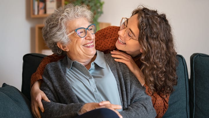 Younger woman embracing senior woman sitting on couch.