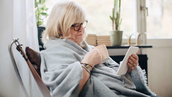 Older woman wrapped in a blanket looking at a tablet while sitting in a chair