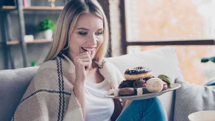 Woman sitting on a couch holding a plate of desserts – including a donut, macarons and other hidden items – looking intently at them and smiling.