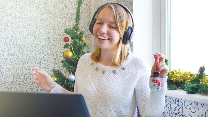 A smiling young woman wearing headphones and looking at a laptop, while holding up a snowman ornament.