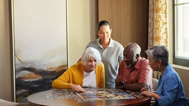 Residents at The Landing at Watercrest Shadow Creek work on a jigsaw puzzle at a round table with a caregiver standing nearby, offering support in a warm, sunlit room.