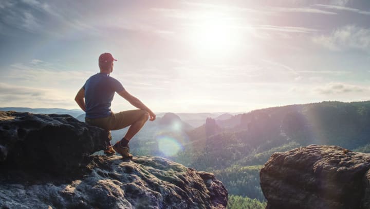 Man sitting at the top of a mountain while looking out onto the surrounding  territory with the sun setting ahead.