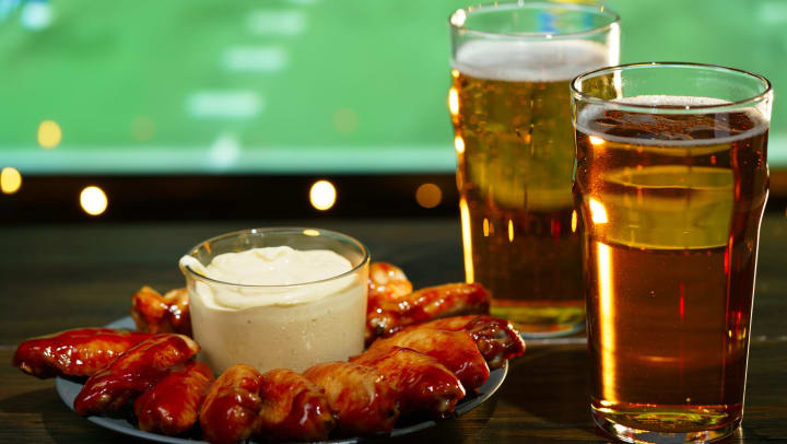 Barbecue wings and two glasses of beer in the foreground with football playing on a TV in the background.