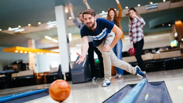 Man rolling a bowling ball down an alley while three people cheer in the background.