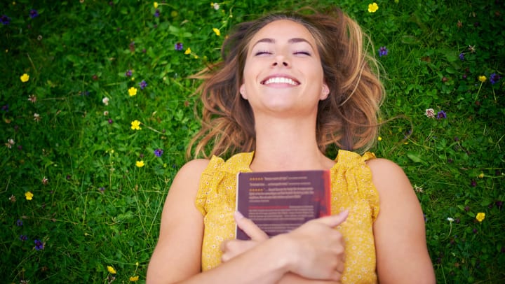 Woman lying in the grass while holding a book to her chest and grinning. 