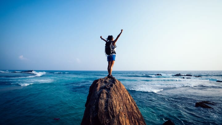 A woman standing on a rock at the edge of the ocean with her arms raised in the air.