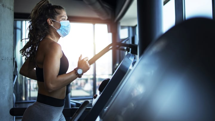 A woman in athletic wear and a medical mask runs on a treadmill.