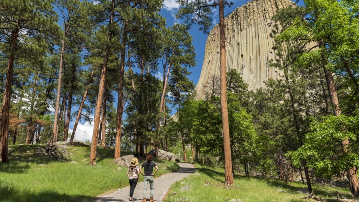 Two people standing on a paved walkway looking up at a massive butte in the background.