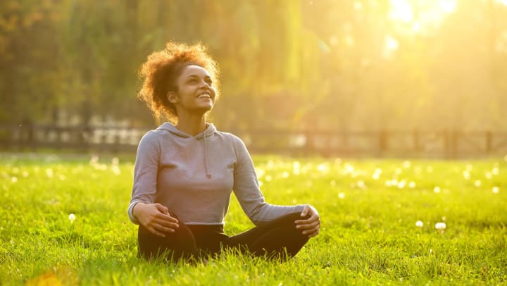 A smiling young woman sitting outdoors in a yoga position with a sunlit backdrop.