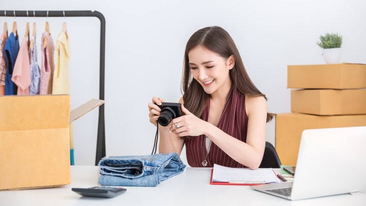A woman sitting at a desk looking at a camera. The desk has an open box, folded jeans, a laptop and other miscellaneous items on it.