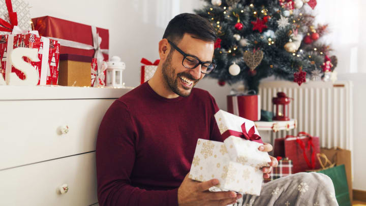 Young man wearing glasses and a maroon sweater opening a wrapped gift while seated on the floor next to a white dresser and Christmas tree