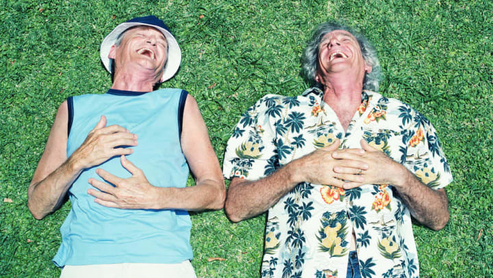 Two older men lying on the grass in the sun and laughing.