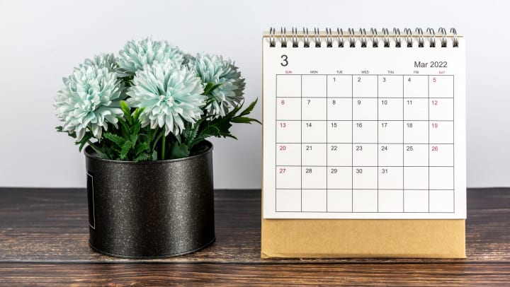 A calendar on a wooden table open to March 2022 sitting next to potted blue flowers. 