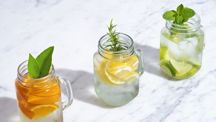 Three Mason jars on a marble countertop filled with citrus fruits and herbs.
