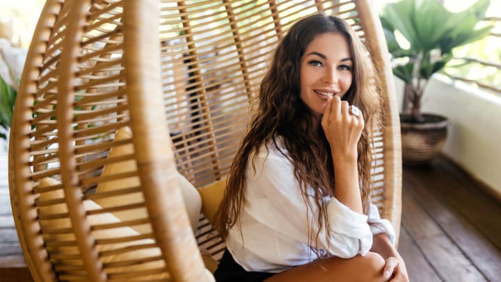 A smiling young woman sits in a round rattan hanging chair. 