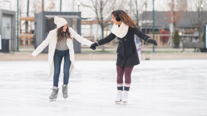 Two women holding hands and ice skating at an outdoor ice rink. 