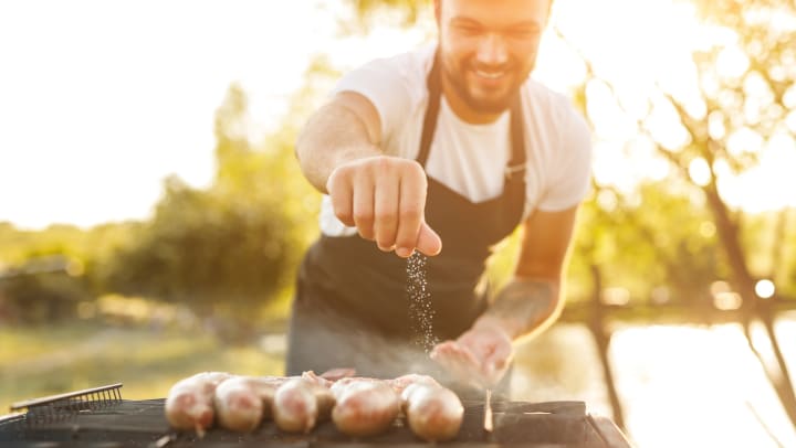Man sprinkling salt on sausages on a grill.