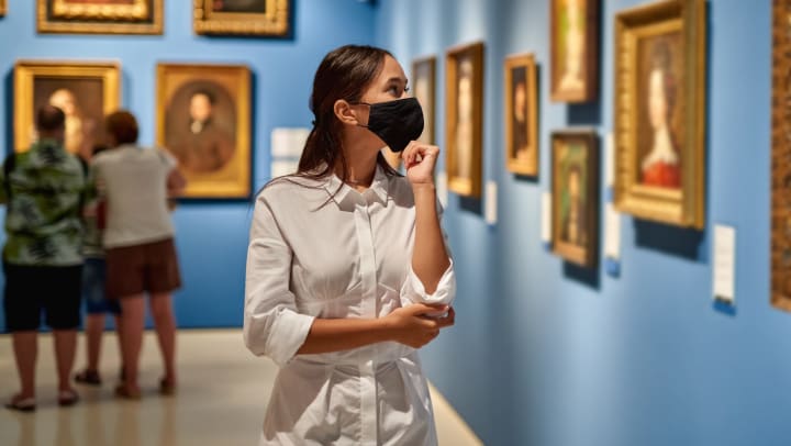 Woman in an art museum with bright blue walls, looking at a painting with her hand to her chin. 