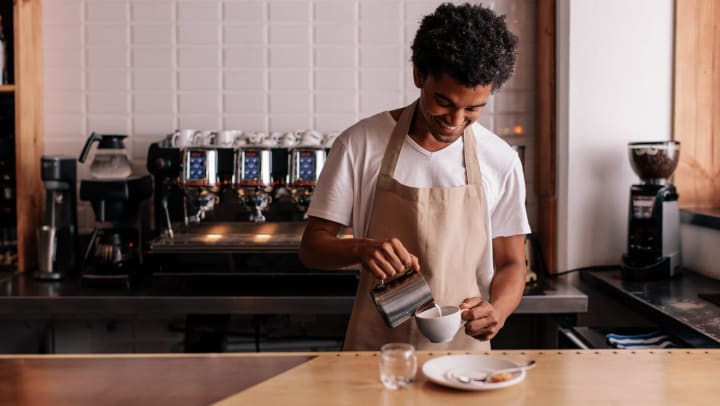 A smiling young man in a coffee shop pouring milk into a cup.