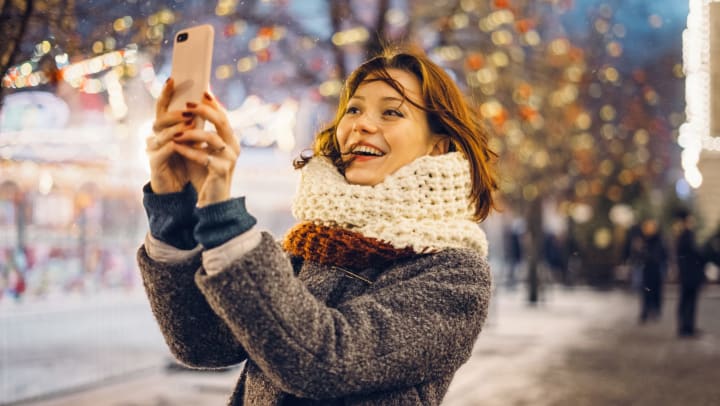 A woman, outside, holding up her phone and smiling. Trees with Christmas lights in the background.  