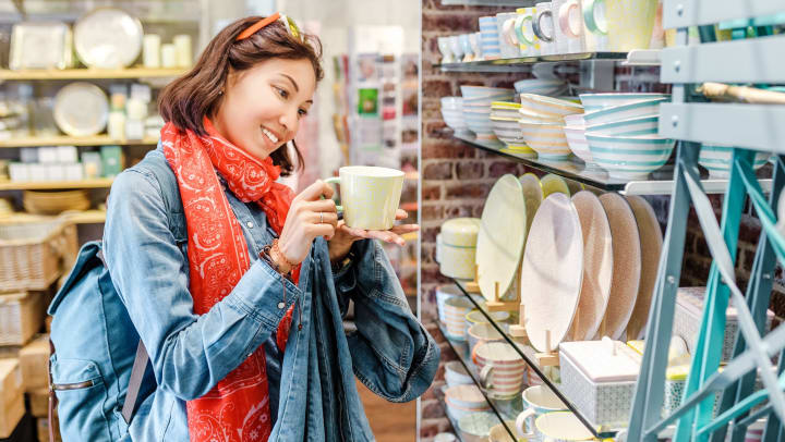 Smiling young woman wearing a denim shirt and red bandana scarf holding a pastel yellow mug in a colorful retail store with kitchenware items