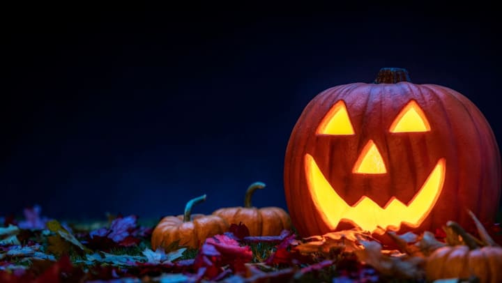 A smiling Jack O’ Lantern sitting in the grass with small pumpkins and fallen leaves at night.