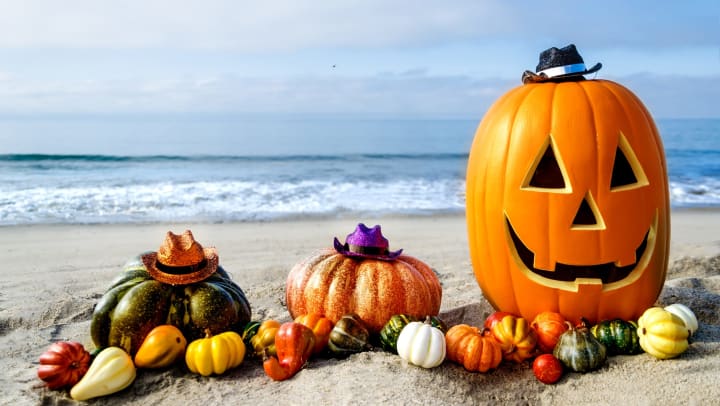 A carved pumpkin and an assortment of multi-colored pumpkins on a sandy beach with ocean waves behind them