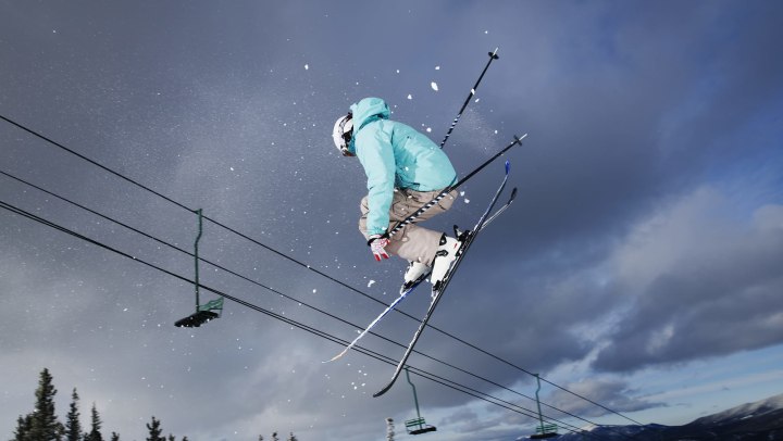 Skier flying off jump with a chairlift and trees in the background.