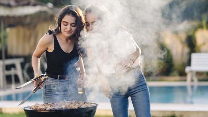 Two women standing over a grill covered in meat looking down. One is holding tongs and a beer.
