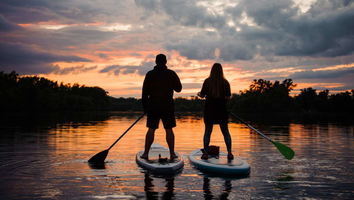 Rear view of a couple of people standing on stand up paddleboards on the river at sunset.