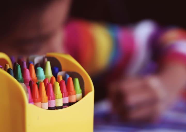 a fuzzy image of a child coloring with an upclose image of a box of crayons in the foreground