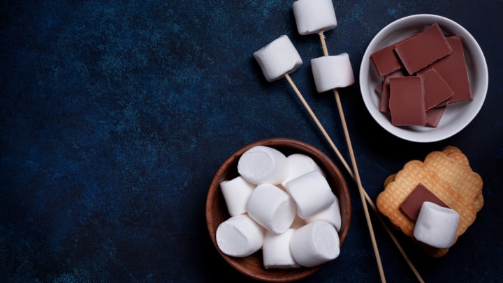 Marshmallows on sticks, chocolate pieces and marshmallows in bowls, and a stack of graham crackers, all on a dark background.