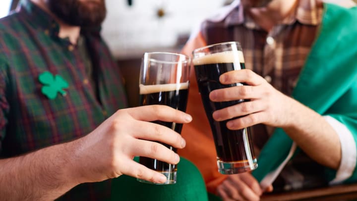 Two men in green smiling and holding glasses full of dark beers up to each other. 