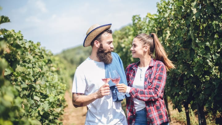 A man and woman couple drinking wine while walking between rows of grape vines.