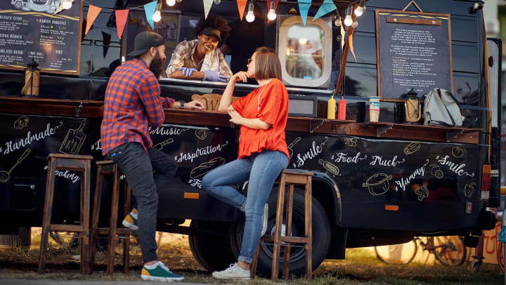 Two people seated in front of a food truck talking to the server inside. 