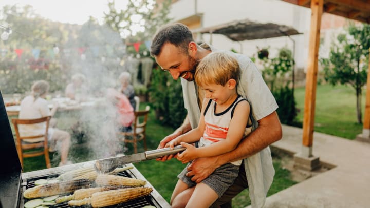 A smiling father showing his boy how to grill corn at a backyard barbecue