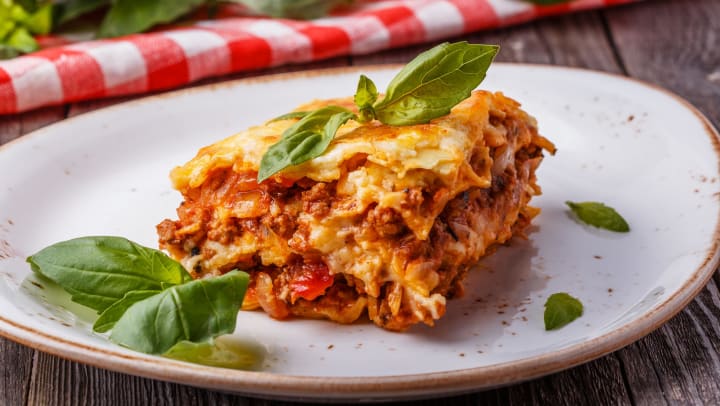 A slice of lasagna on a white plate with a basil on top of it with red checkered fabric and more basil out of focus in the background. 