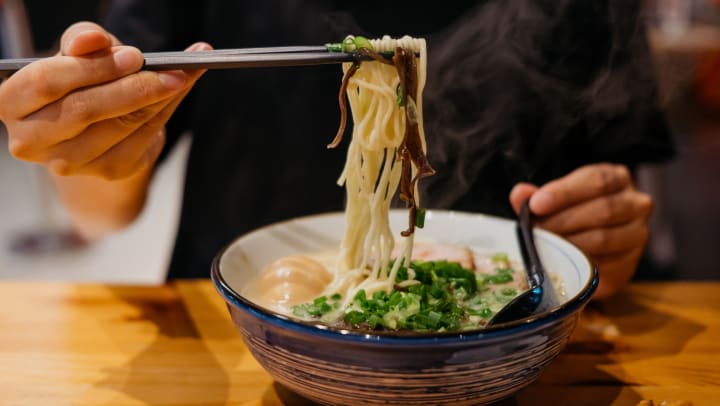 Close up of a person’s hand holding chopsticks as they lift noodles from a bowl of ramen. 