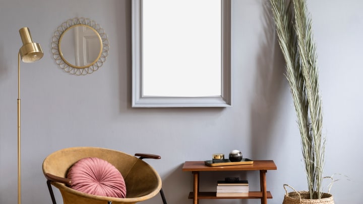 Interior with honey yellow armchair, gray mockup frame, palm, gold mirror and lamp, as well as a retro small table with elegant accessories and books.