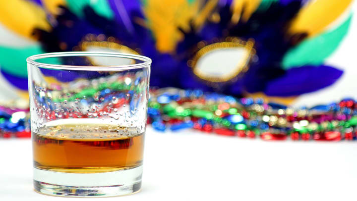 A drink full of amber liquid in front of blurry mardi gras beads and a mask. 